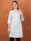 Model wearing an Eelika white cotton kurti with blue floral print, round neck, and 3/4 sleeves against a brown wall.