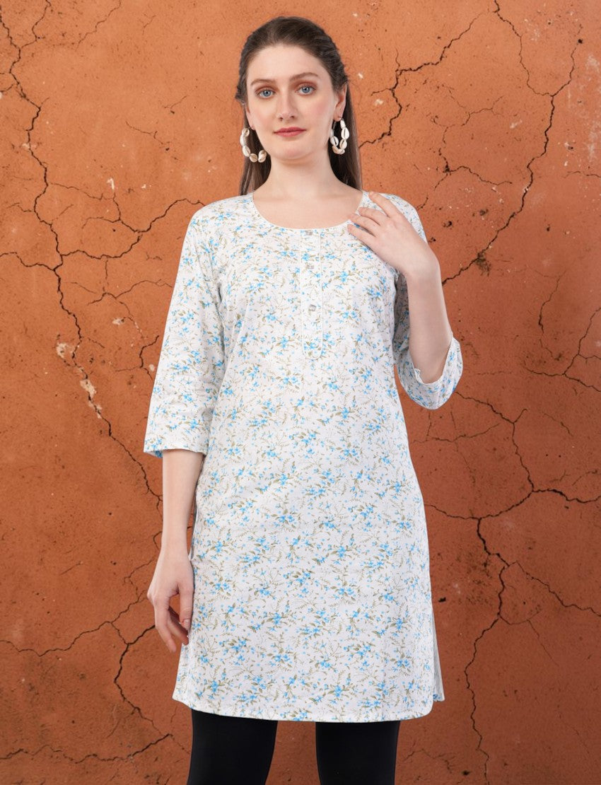 Model wearing an Eelika white cotton kurti with blue floral print, round neck, and 3/4 sleeves against a brown wall.